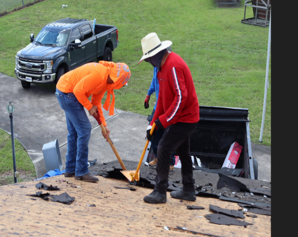 Two men in work clothes removing shingles from a roof. A pickup truck and green car are in the background.