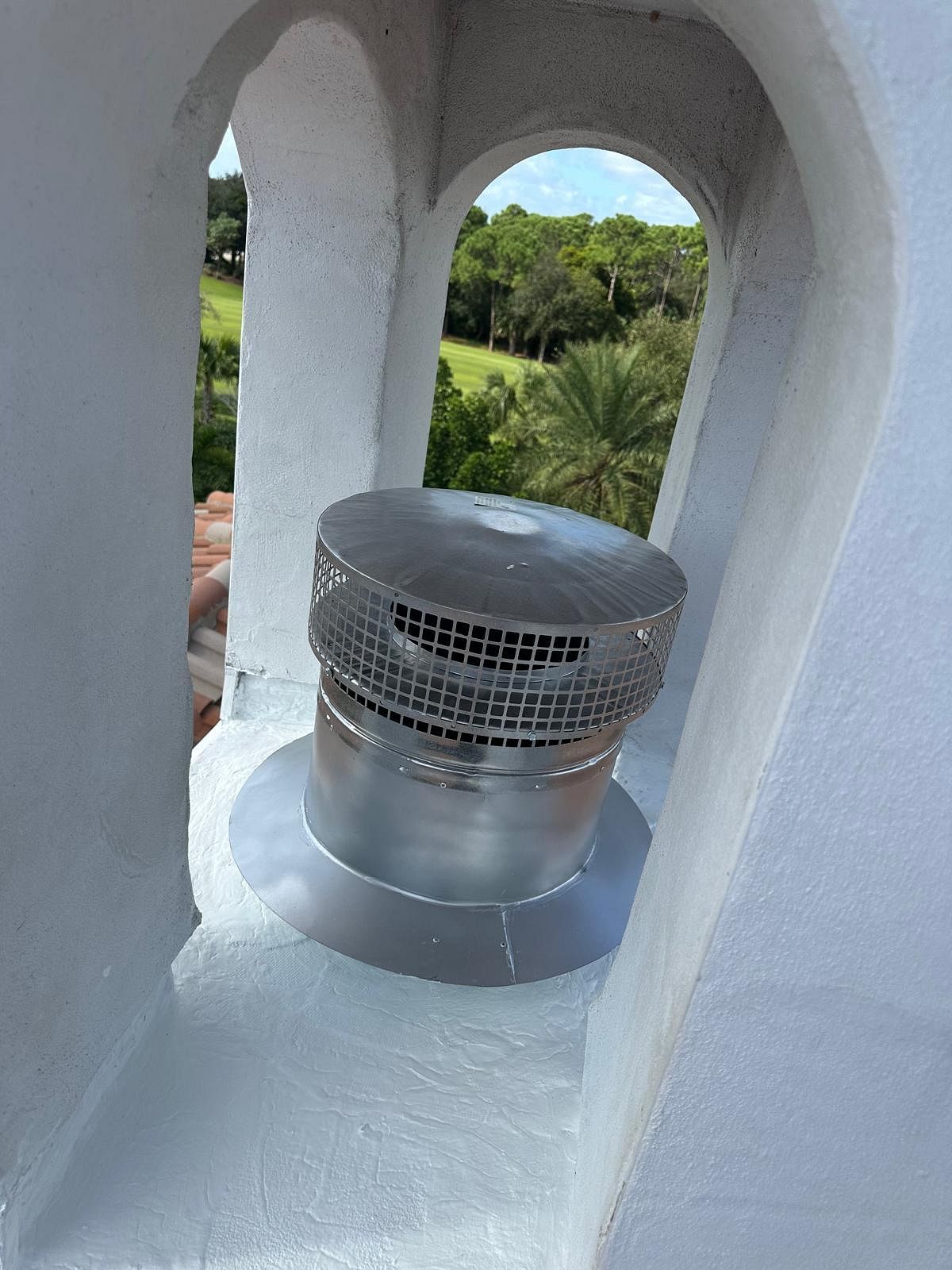 Metal vent pipe on a white building, framed by an archway, with a view of trees and sky.