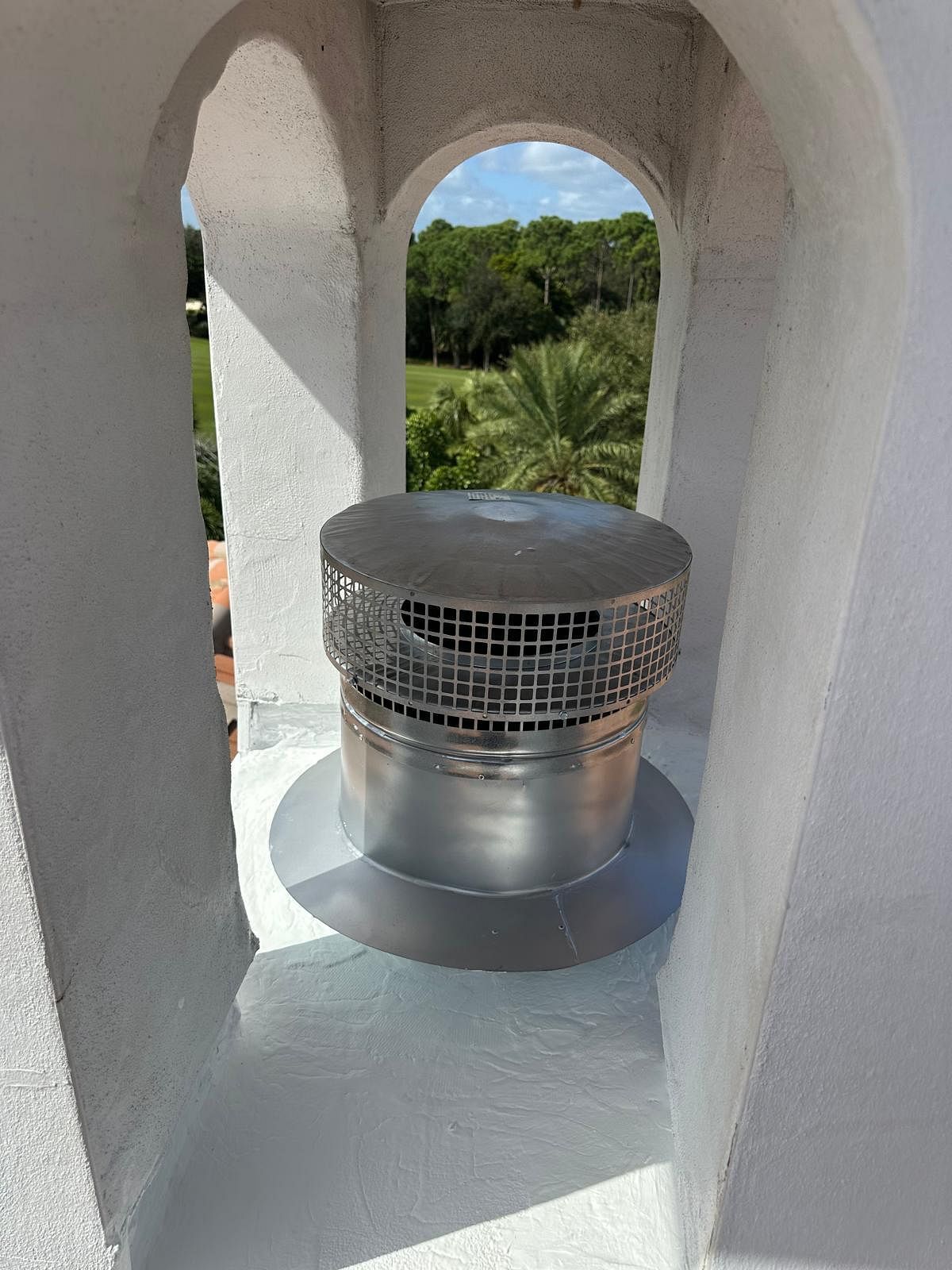 Ventilation cap on a white building's roof, framed by an archway, view of green trees and sky beyond.
