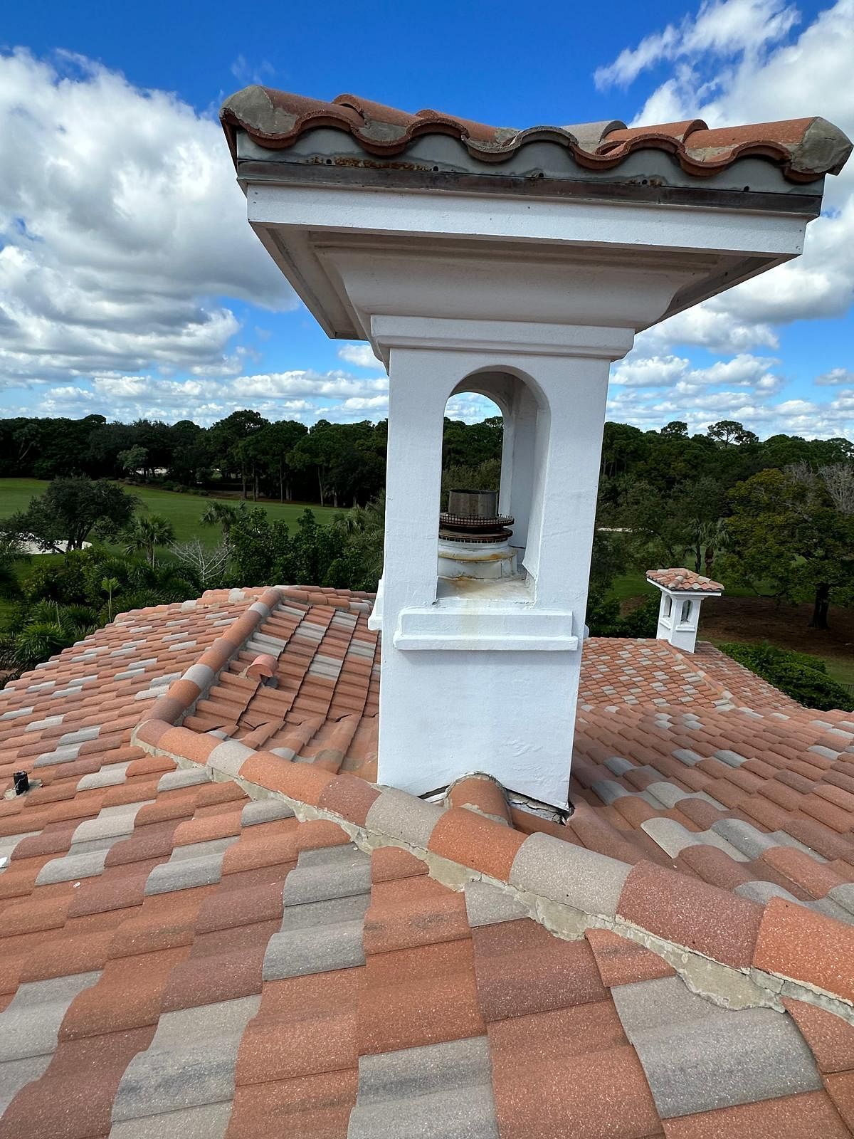 A chimney with red-brown roof tiles, white trim, and a view of green trees and blue sky.