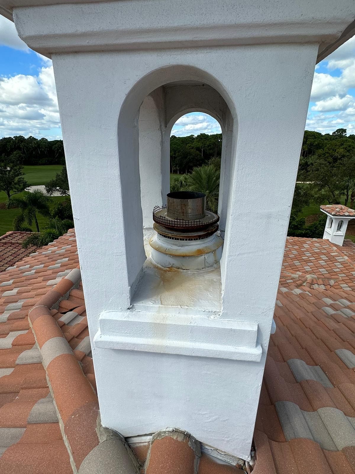 White chimney with arched opening, rusty metal cap, on a red tile roof, overlooking green trees.