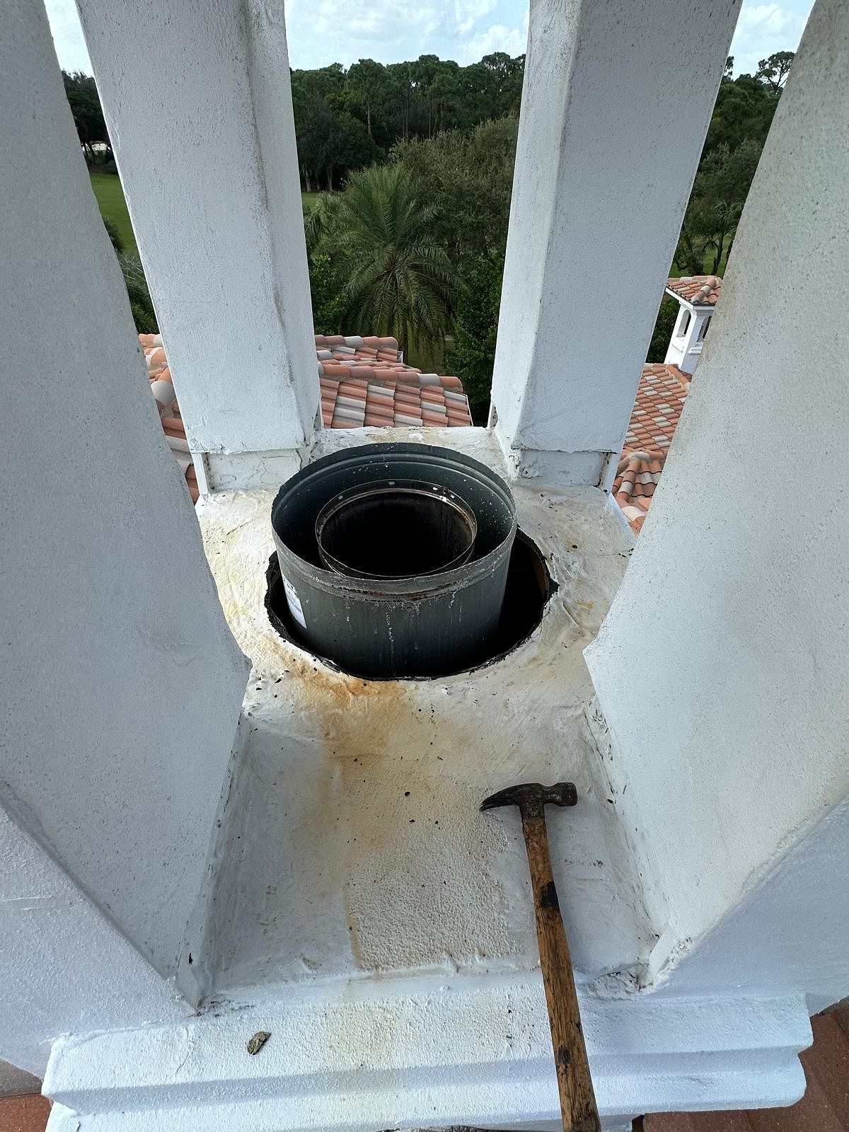 Chimney with metal flue and hammer on a concrete structure, surrounded by roof tiles and greenery.
