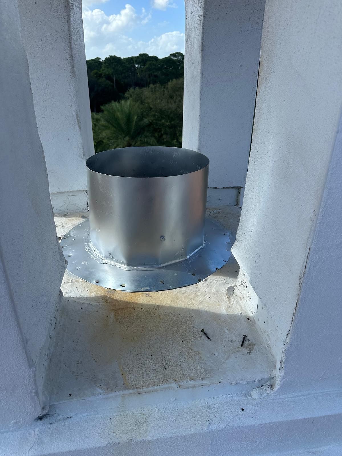 Metal vent pipe on a white roof, with a view of trees and sky in the background.
