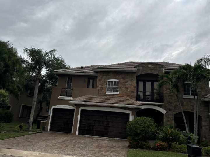Large two-story house with brown garage doors, stone facade, and palm trees under a cloudy sky.