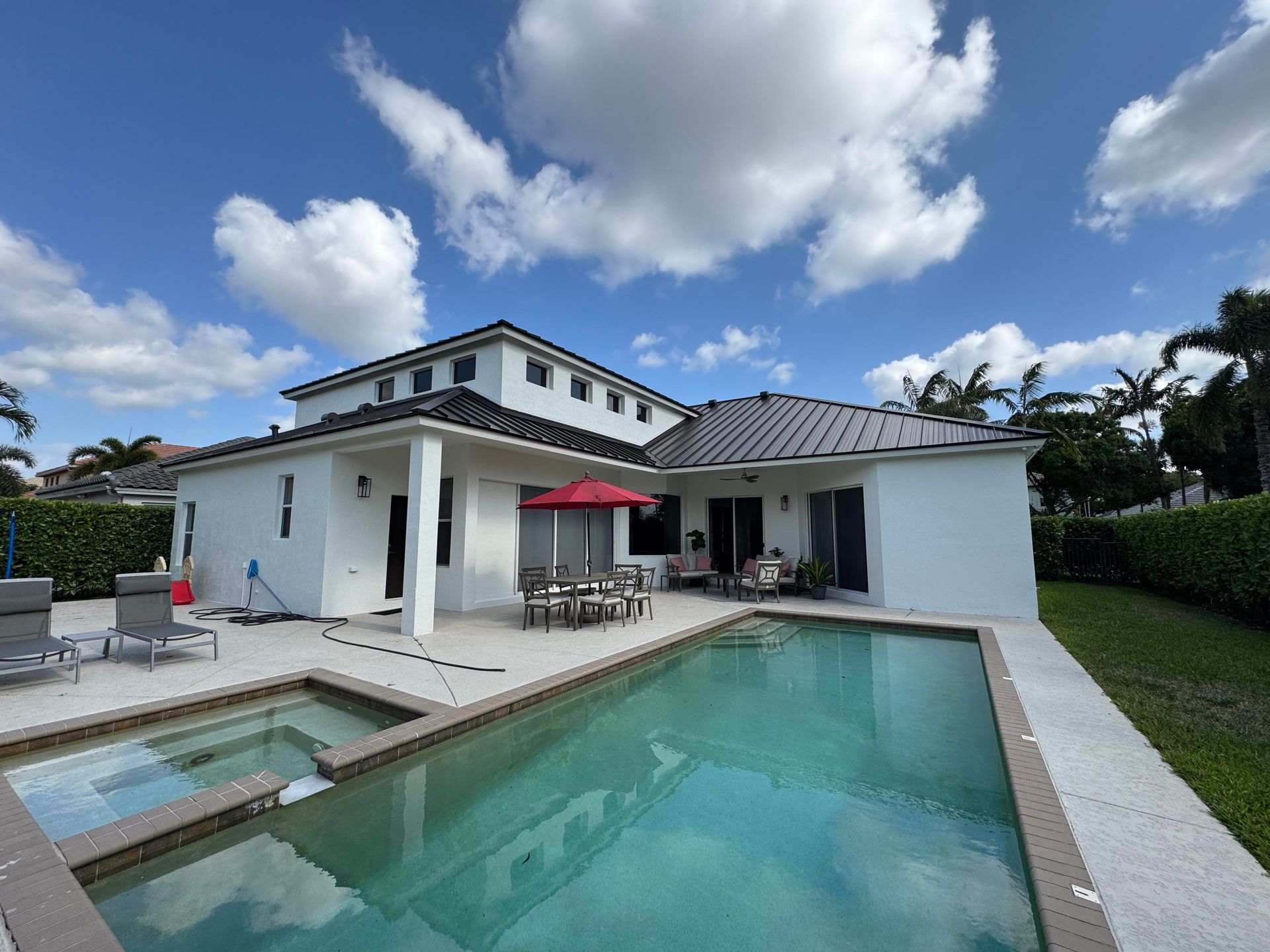 White house with pool, patio, and red umbrella under a blue sky with fluffy white clouds.