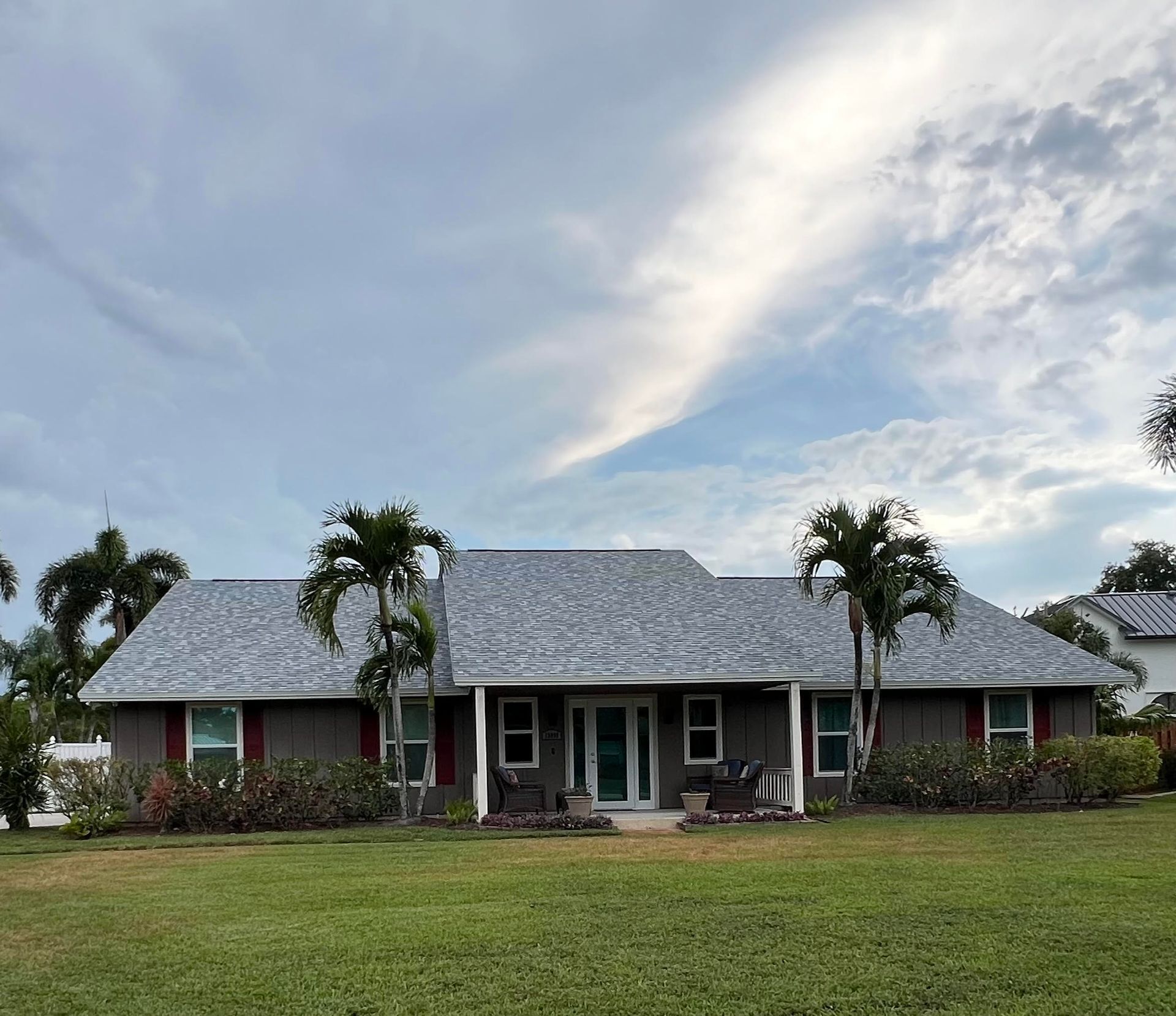 A brown house with a gray roof, palm trees, and a green lawn under a cloudy sky.