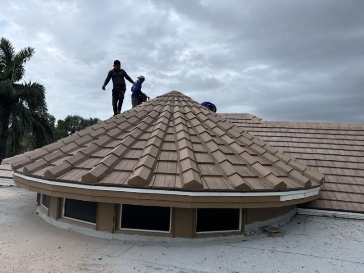 Roofers installing brown tile shingles on a round-roofed building under an overcast sky.