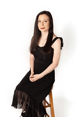 Woman in a black dress seated on a wooden stool against a white background