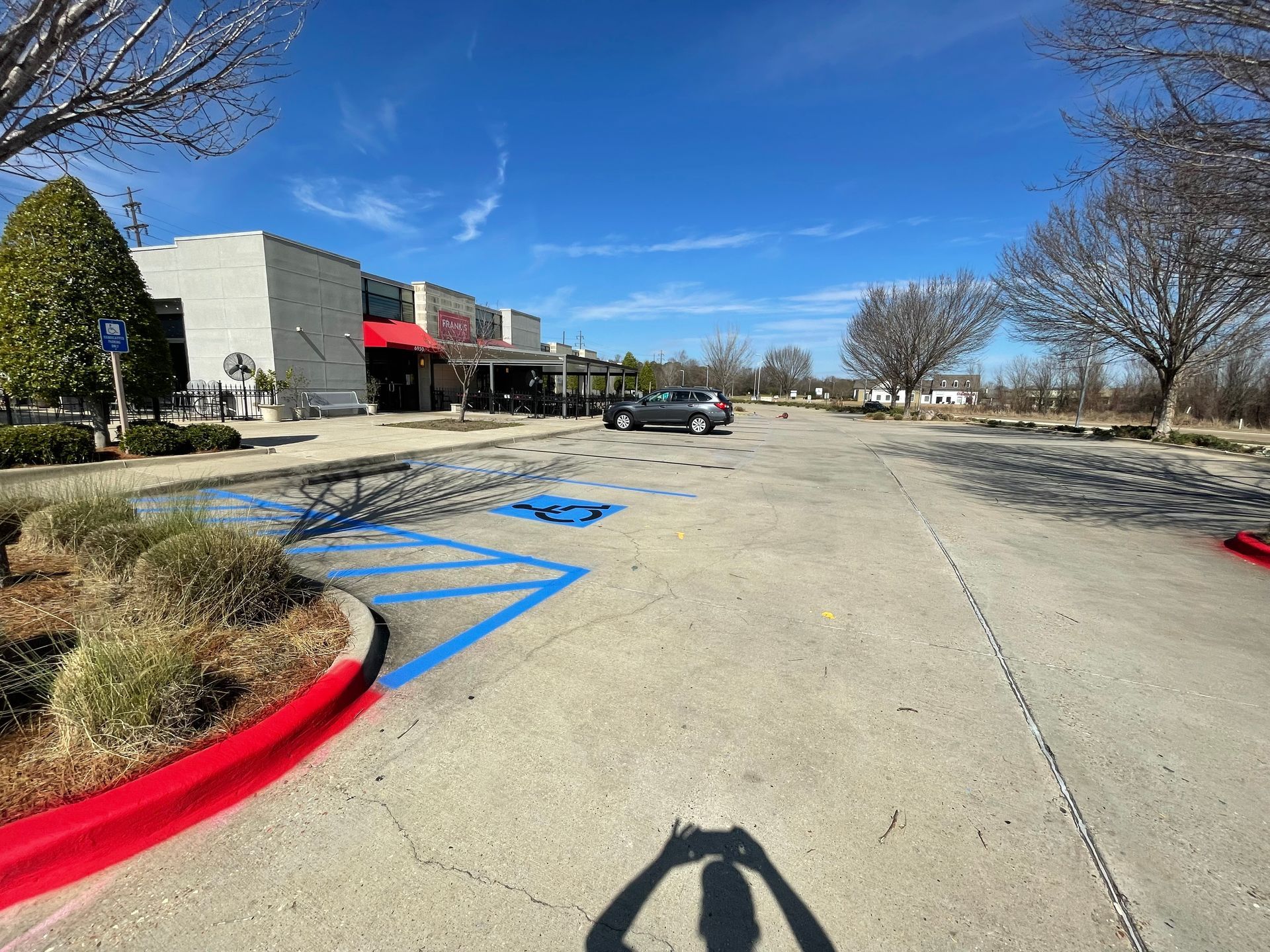 Parking lot in front of businesses on a sunny day. A gray car is parked. Person taking photo casts a shadow.