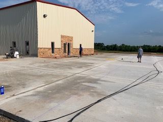Workers outside a tan metal building with a brick facade, on a concrete parking area on a sunny day.