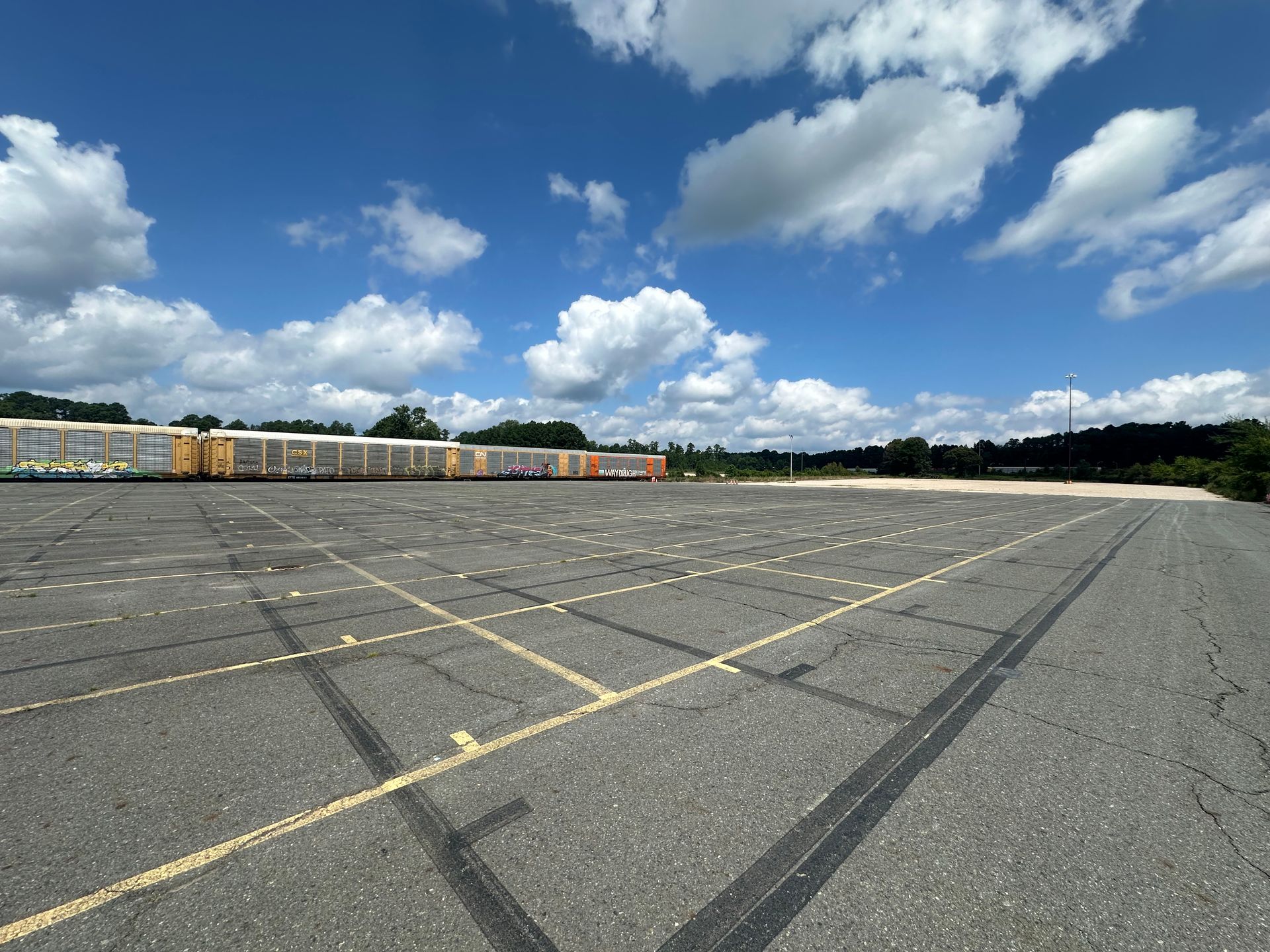 Wide shot of a large empty parking lot under a blue sky with fluffy clouds; a train is in the distance.