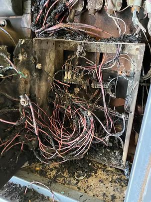 Burnt electrical panel with melted wires and charred components, likely due to fire damage.