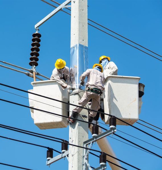 Linemen in two bucket trucks working on power lines attached to a utility pole against a blue sky.
