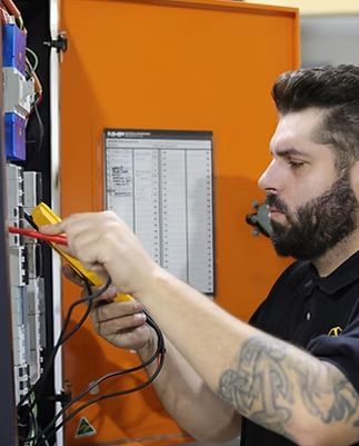 Man using a multimeter to test electrical panel. Orange cabinet, black cords, and visible tattoo.