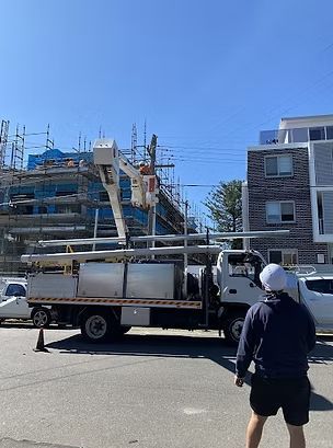 A utility truck with a lift arm reaching towards power lines, worker standing nearby. Construction site backdrop.