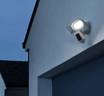 White security light and camera mounted on a blue-gray building exterior, light illuminated.