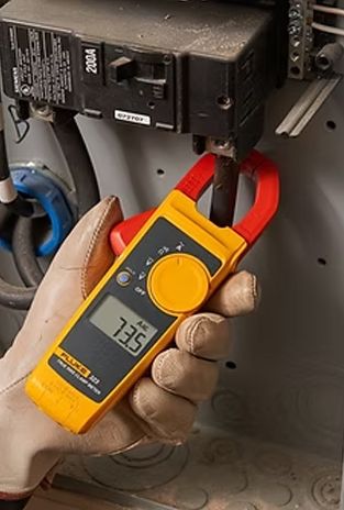A gloved hand uses a yellow clamp meter on electrical wiring, reading 73.5.
