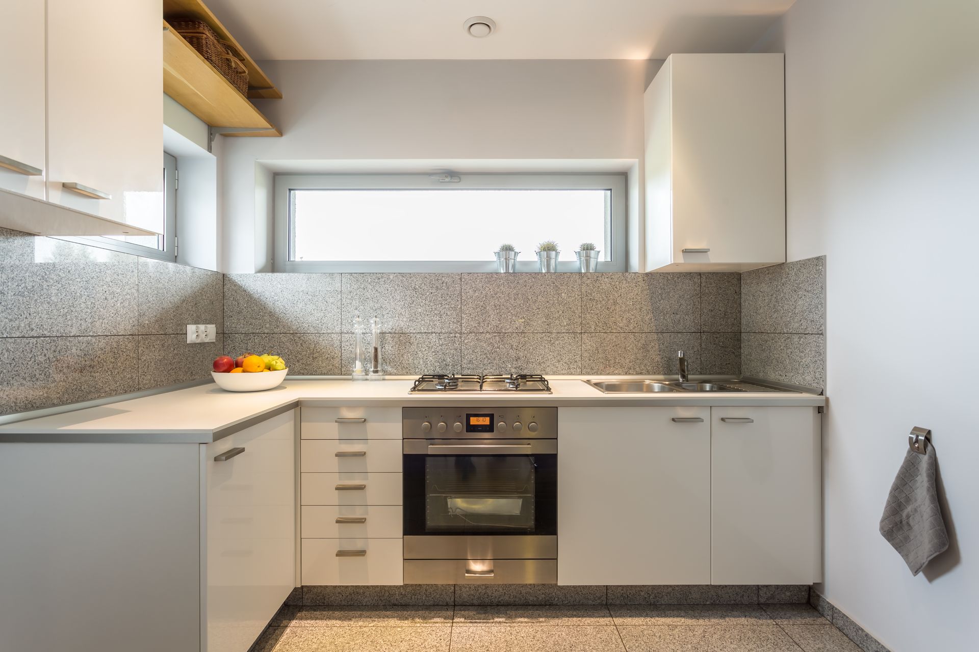 Modern white kitchen with built-in oven, cooktop, and stainless steel backsplash. Overhead cabinets and under-cabinet lighting.
