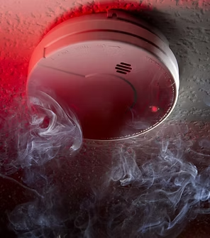 Smoke billowing from a smoke detector mounted on a ceiling; red light is on.