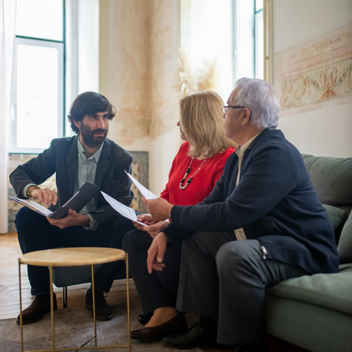 Man in suit consults with couple on a couch, reviewing documents in an office setting.
