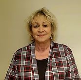 A person with blonde hair wearing a red, white, and black plaid cardigan over a black shirt against a plain wall.