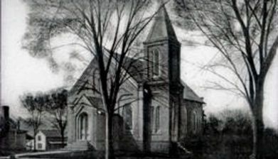 A historic brick church with a tall steeple stands nestled between several large, bare trees in a black-and-white photo.