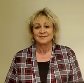 A person with light blonde hair wearing a black shirt and a red, white, and black plaid cardigan against a plain wall.