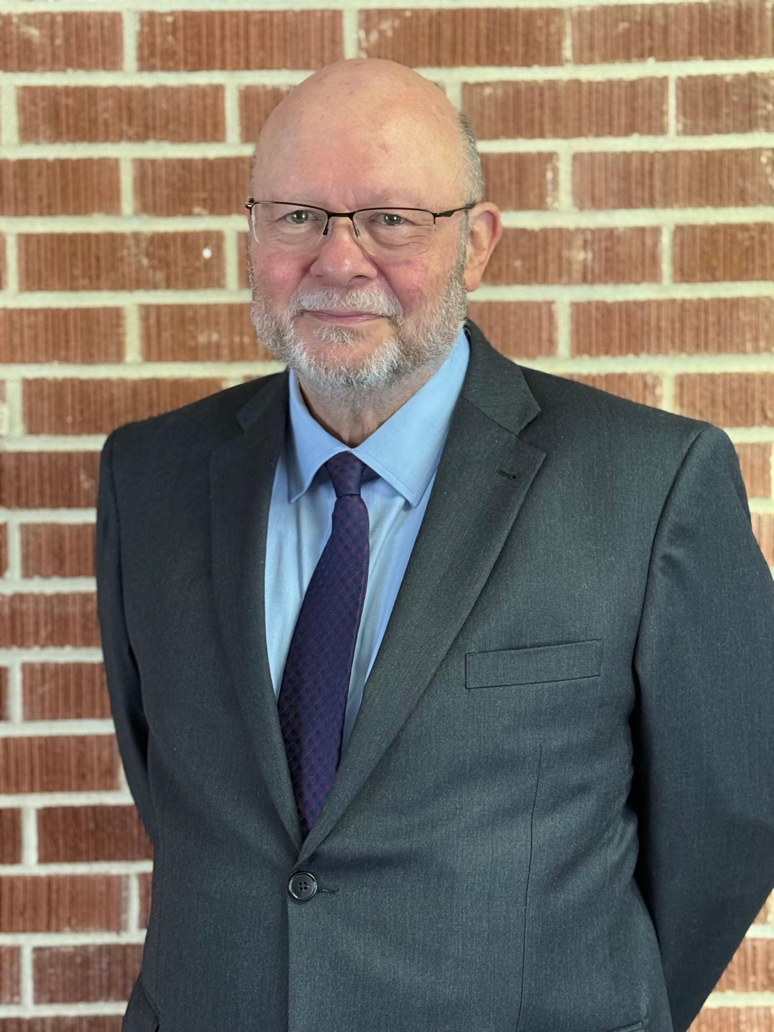 A person wearing a suit, button-down shirt, and tie stands against a brick wall, looking forward.