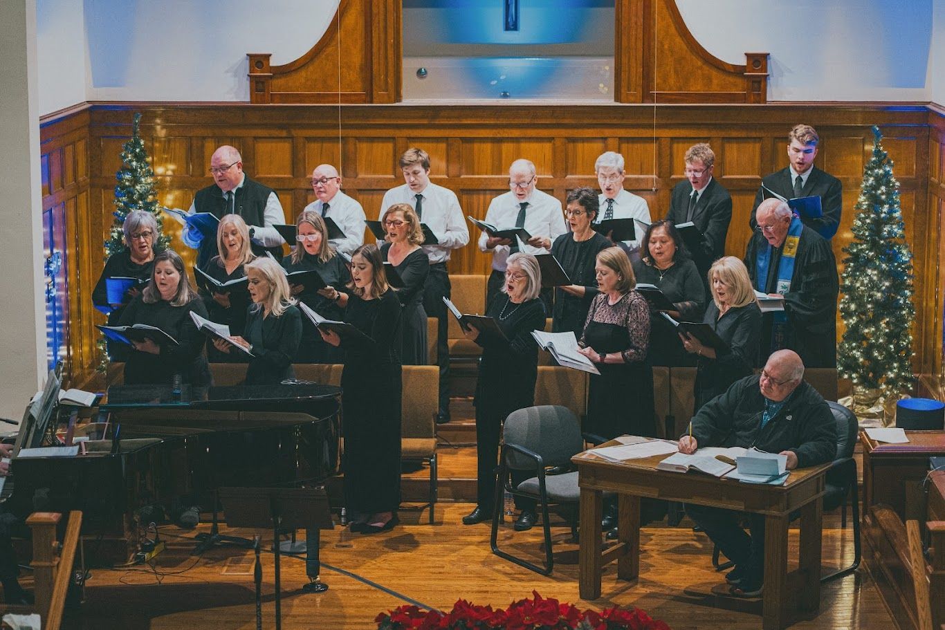 A choir stands in rows on a stage in front of wood paneling and two lit Christmas trees, singing from sheet music.