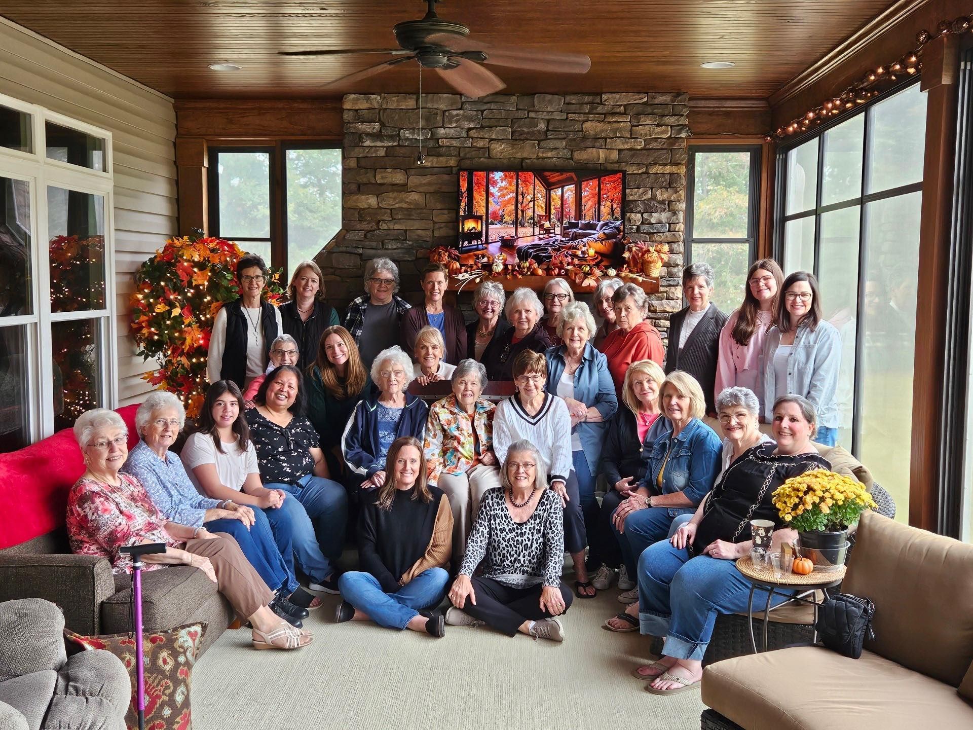 A large group of people poses for a photo on a screened-in porch with a stone fireplace and fall decor.