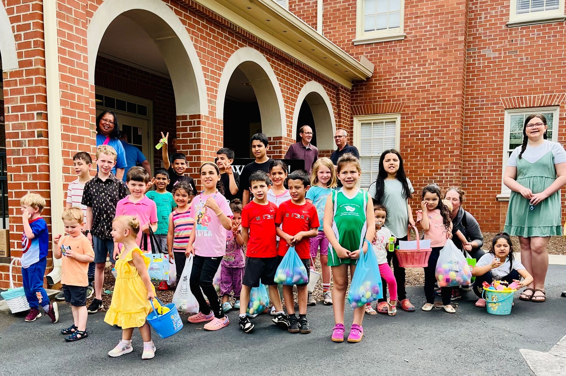 A group of children and adults standing outside a brick building, many holding colorful buckets during an Easter egg hunt.