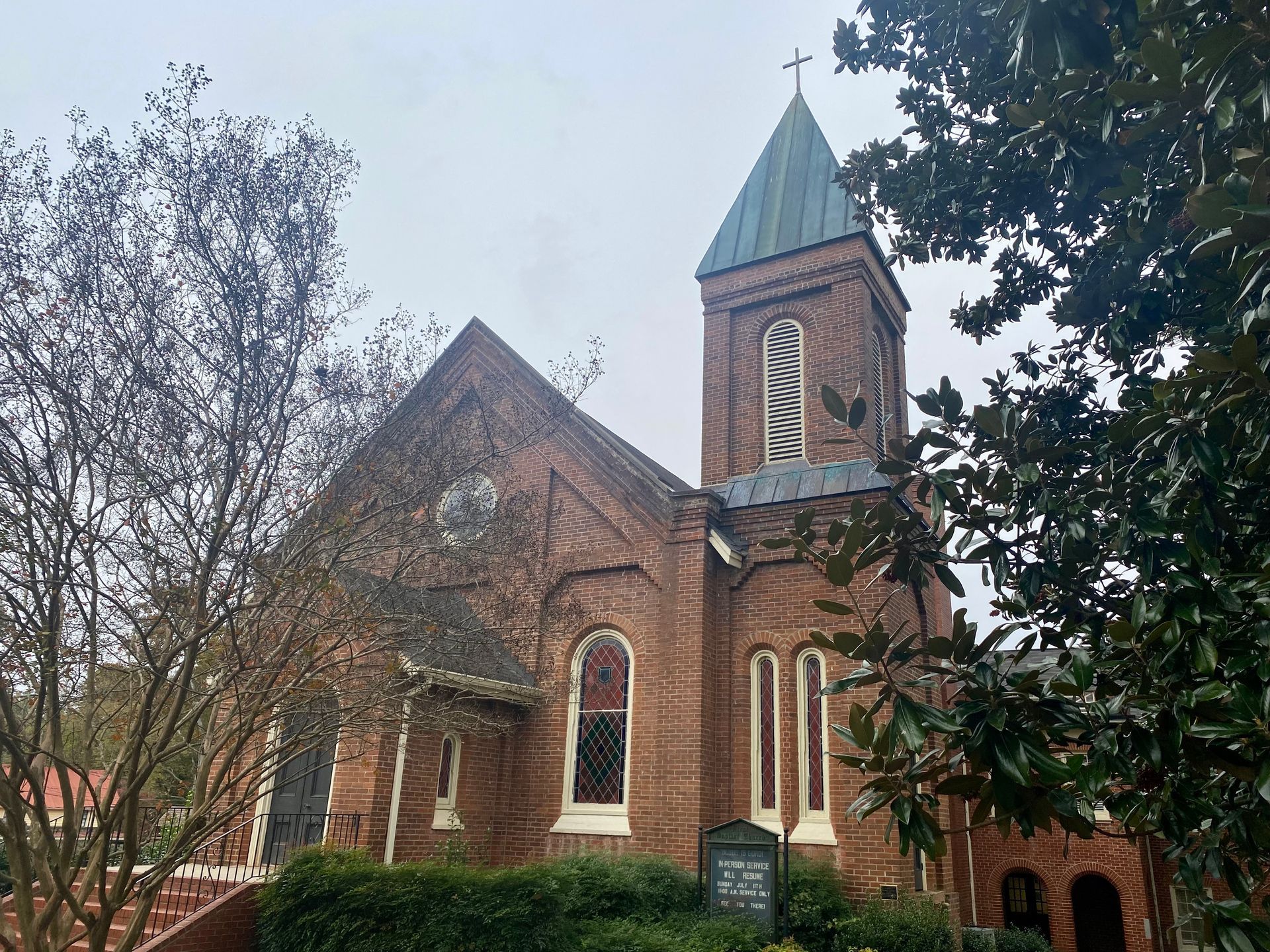 A historic brick church with a tall steeple, stained glass windows, and lush trees in front under an overcast sky.