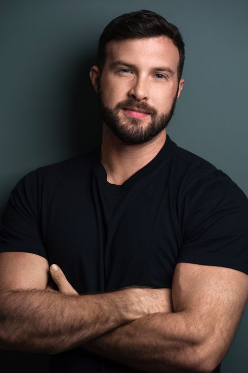 A person with a beard and dark hair wearing a black t-shirt, arms crossed, standing against a dark teal background.