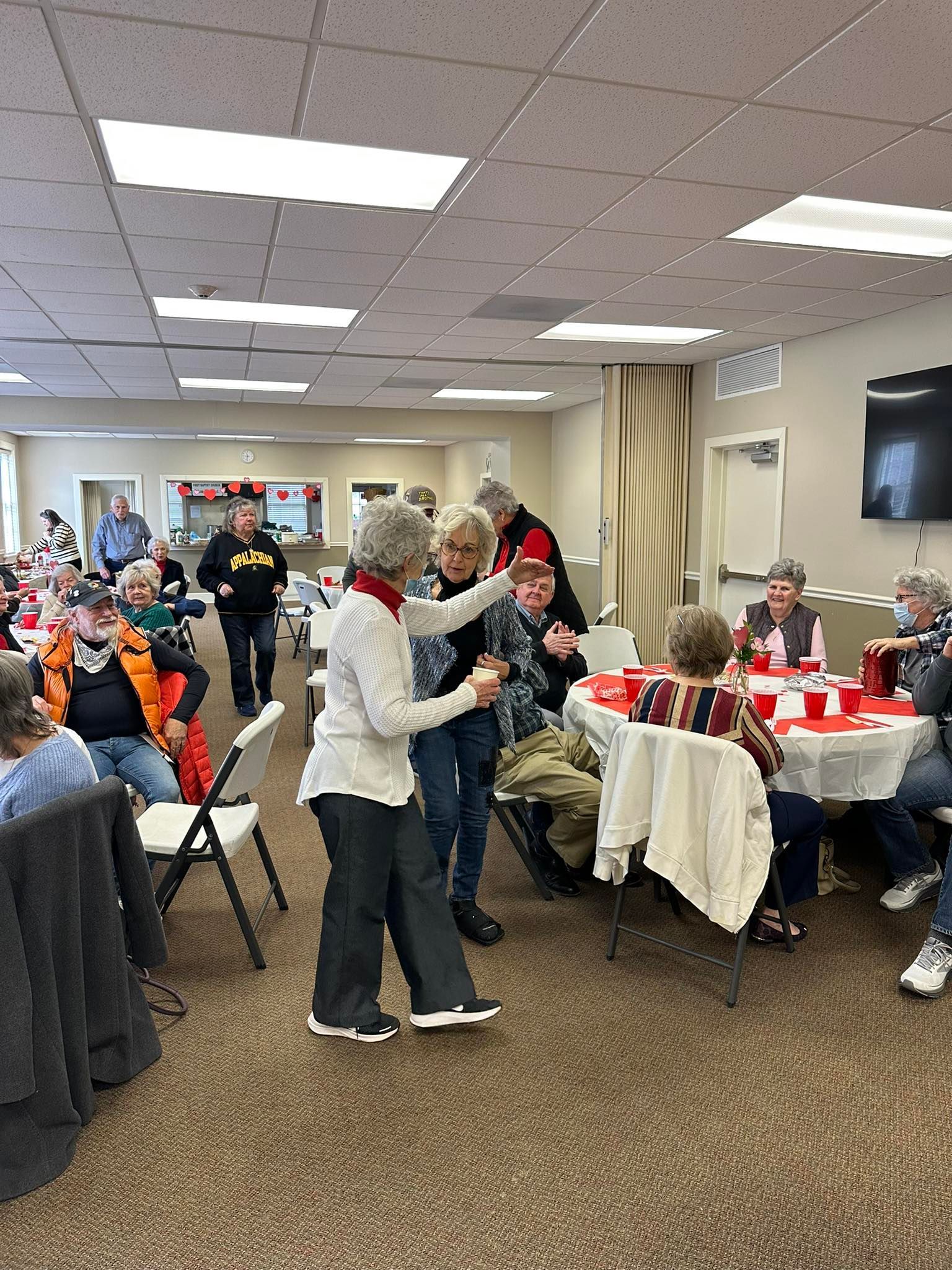 People gather in a community hall for a social event, with some standing to talk while others sit at round red tables.