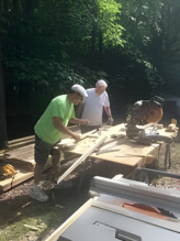 Three people working with wood on a large outdoor table in a wooded area.