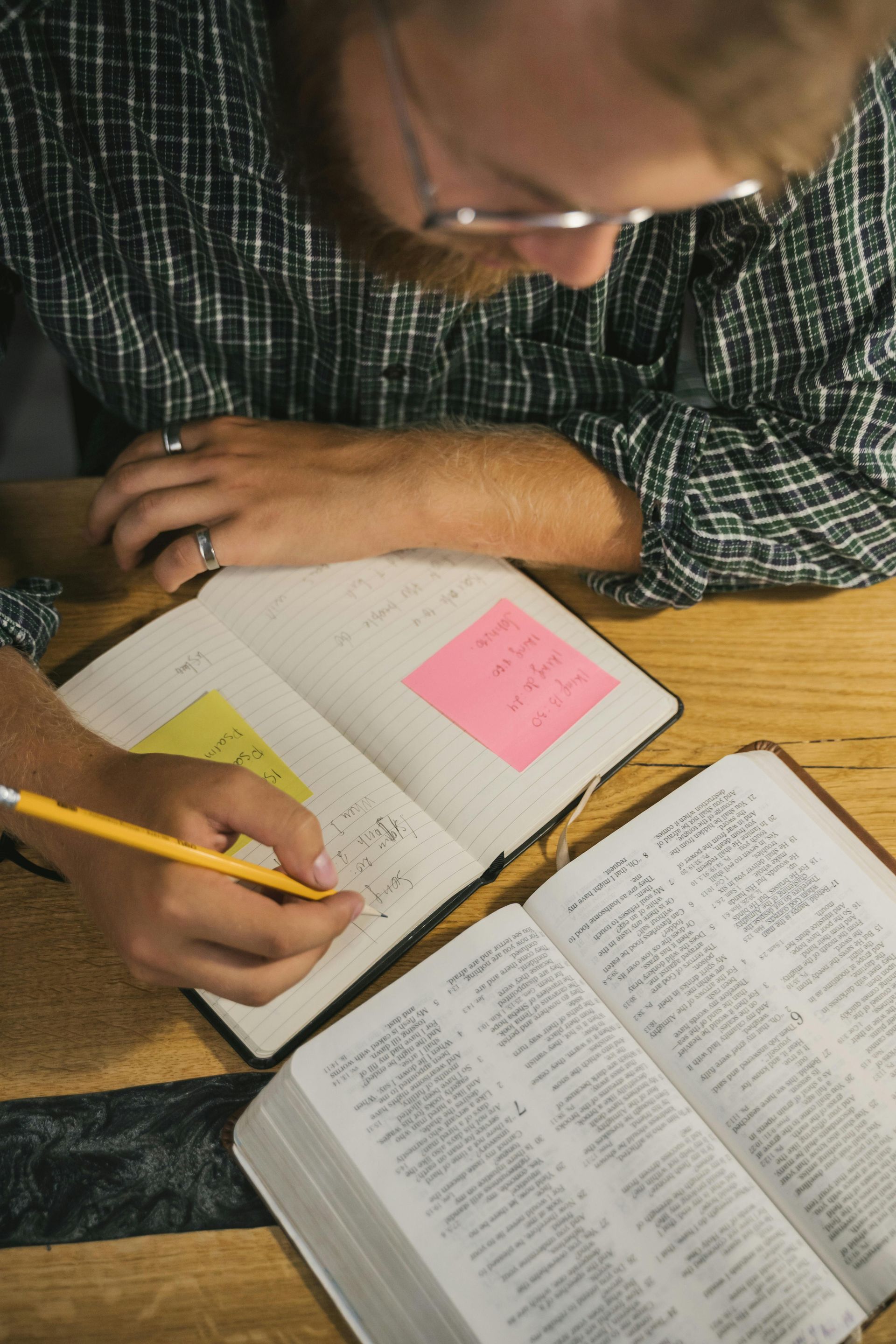 A person in a plaid shirt sits at a wooden table, writing in a notebook next to an open book.