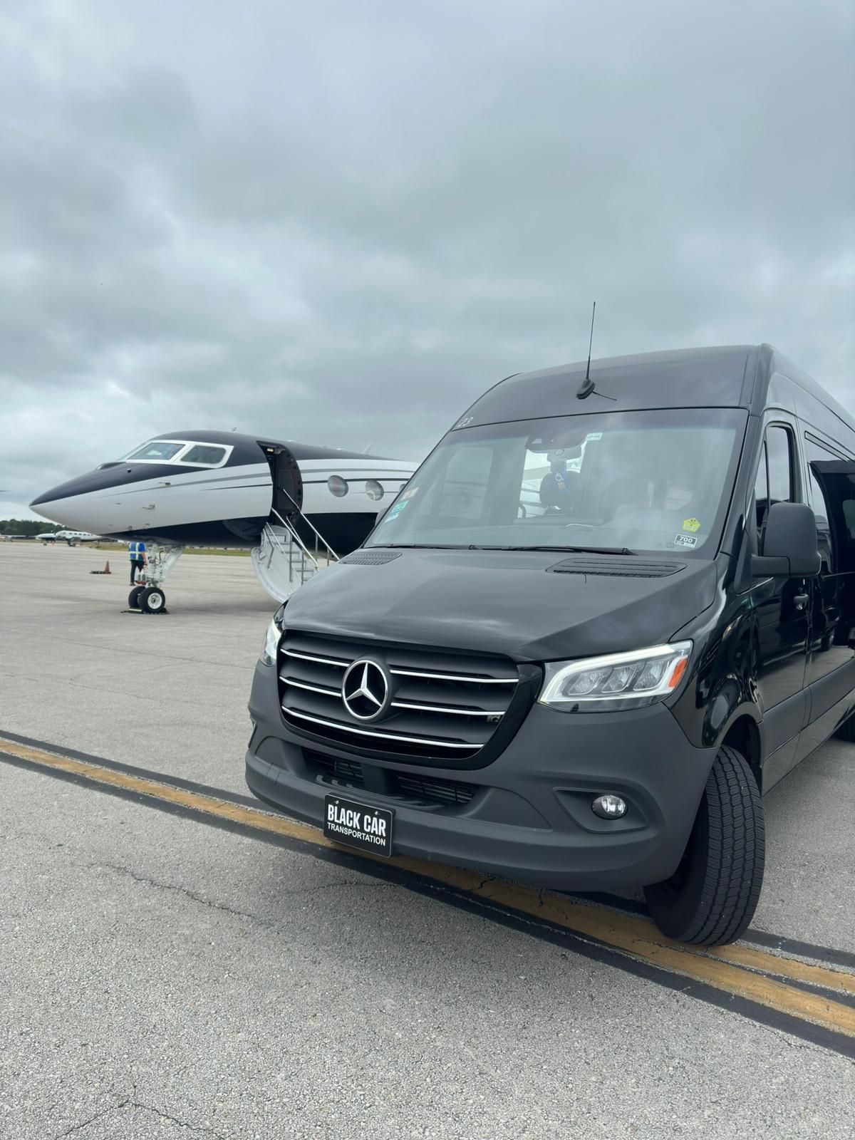 A black mercedes sprinter van is parked on the tarmac next to an airplane.