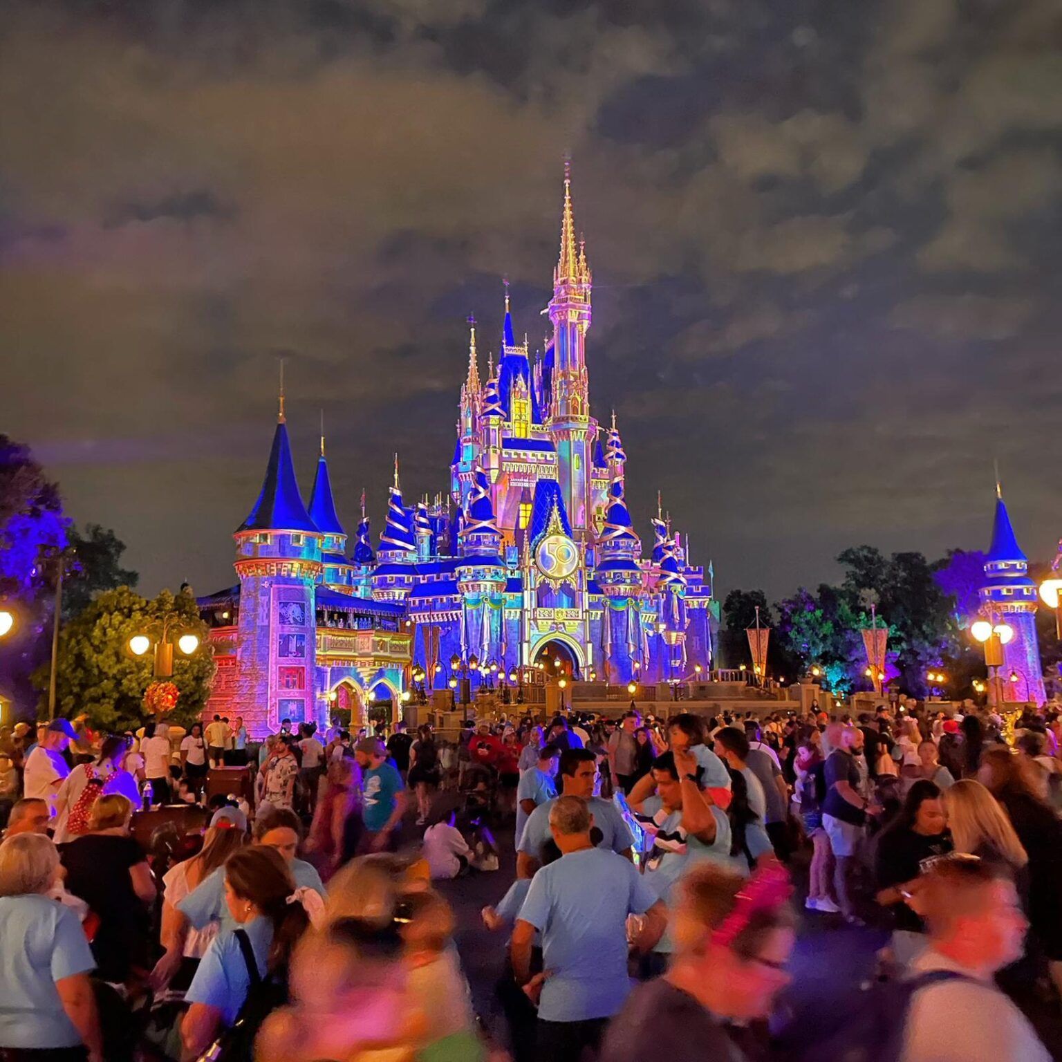 A crowd of people are gathered in front of the disney castle at night