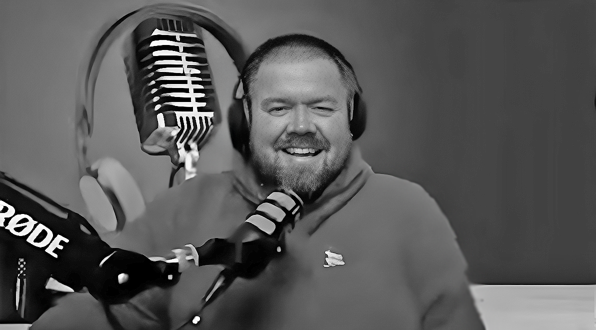 A man wearing headphones and a microphone is smiling in a black and white photo.