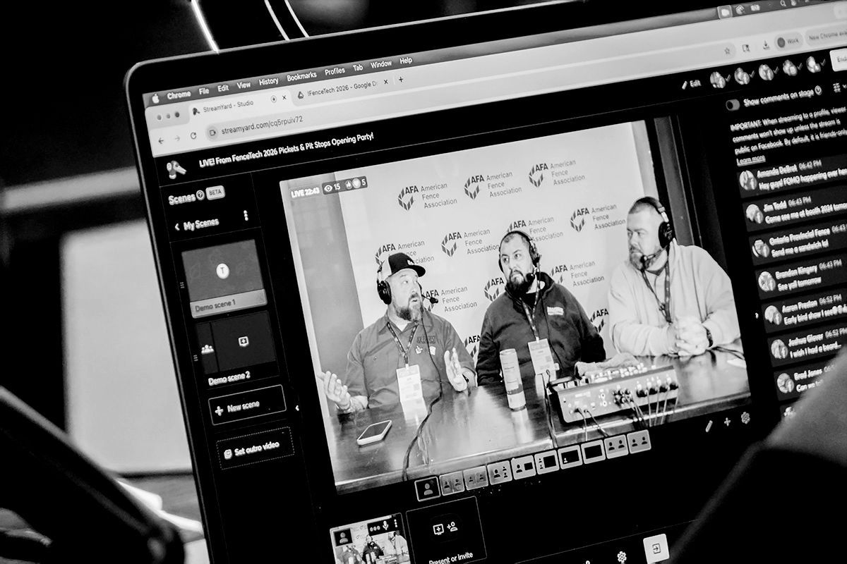 Laptop screen displays a live stream. Three men with microphones sit at a table in front of a backdrop with logos.