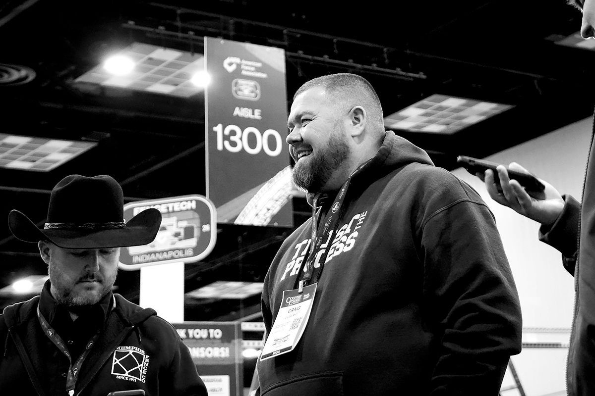 Man smiling with a badge at a convention, another man in a cowboy hat stands nearby.