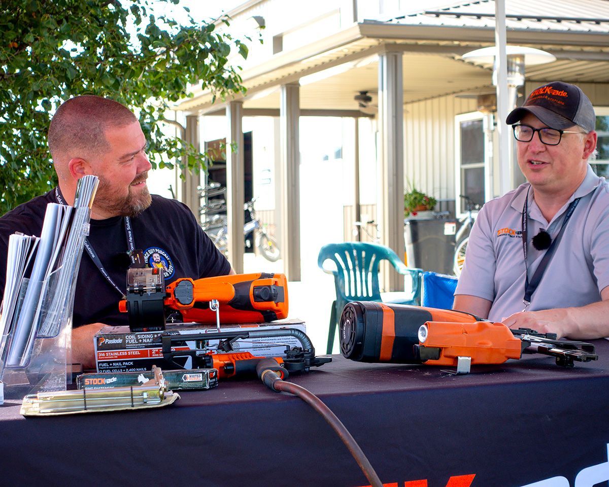 Two men at a table with orange power tools, outdoors, discussing equipment.