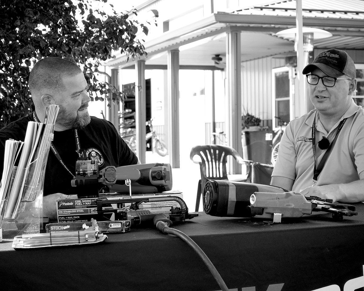 Two men seated at a table with equipment. One bearded man looks towards the other, who is speaking, wearing glasses and a cap.