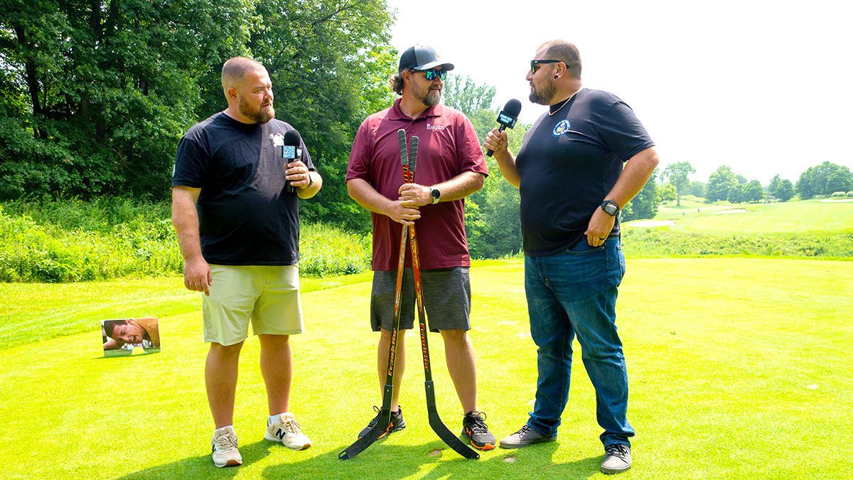 Three men on a grassy field with hockey sticks. One is being interviewed. Green grass, sunny day.