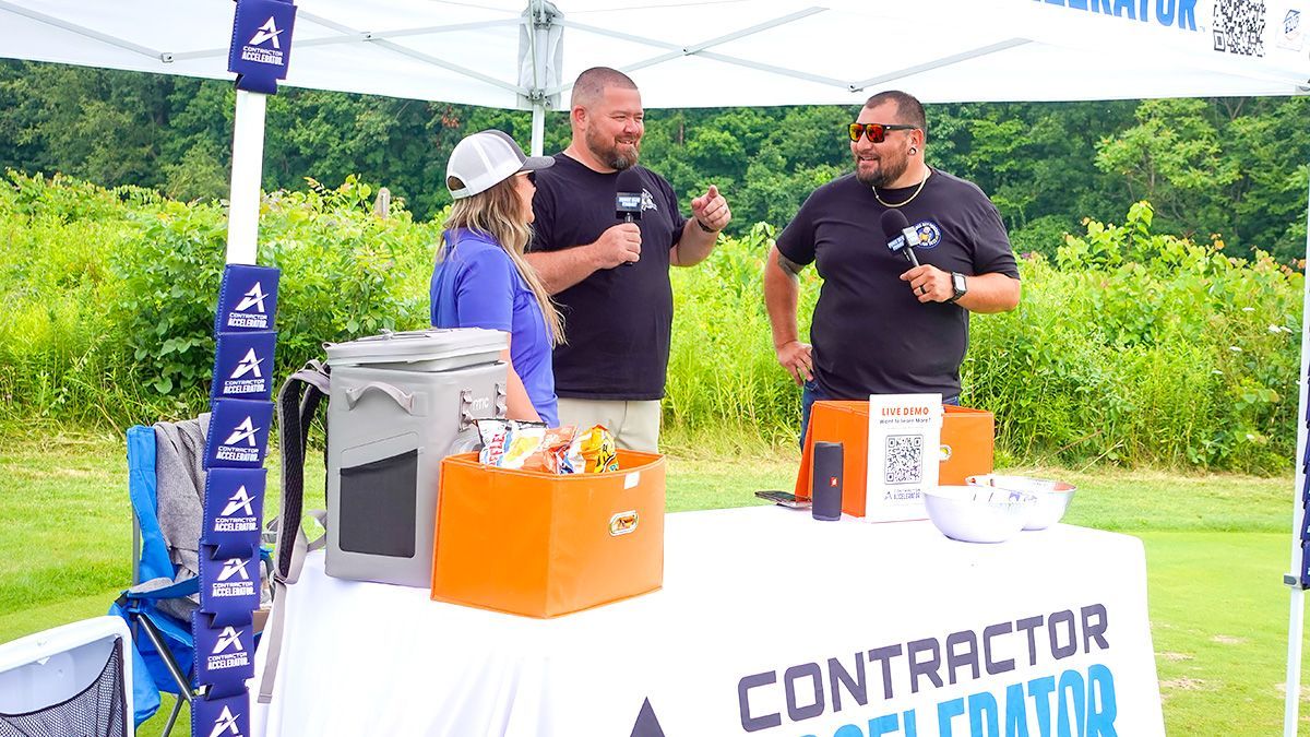 Three people at a Contractor Accelerator booth outdoors. They are smiling and talking, with a cooler and snacks visible.