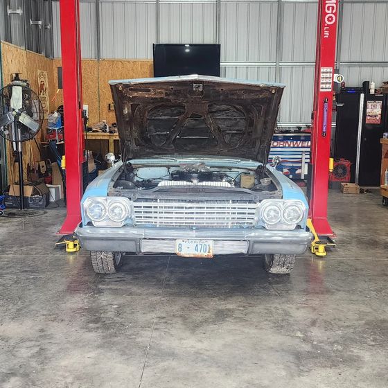 Classic car on a lift in an auto shop, hood open for repair, with red posts and workshop tools around it