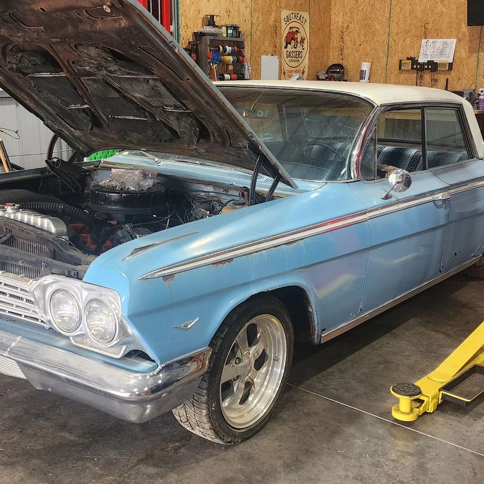 Blue classic car in a garage with hood open, viewed from the front-left side.