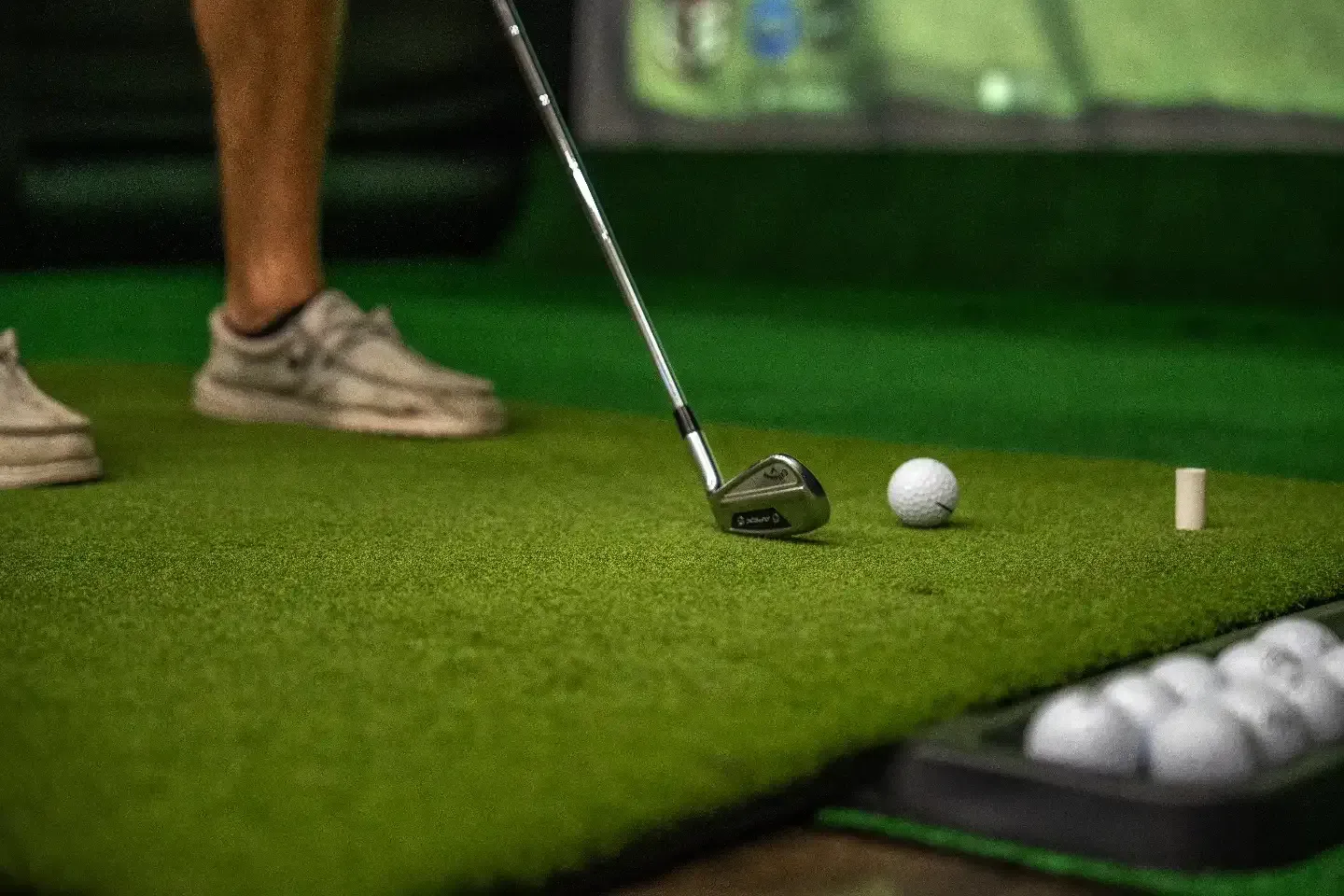 Golfer addressing a ball on a green indoor putting mat, with tees and golf balls nearby