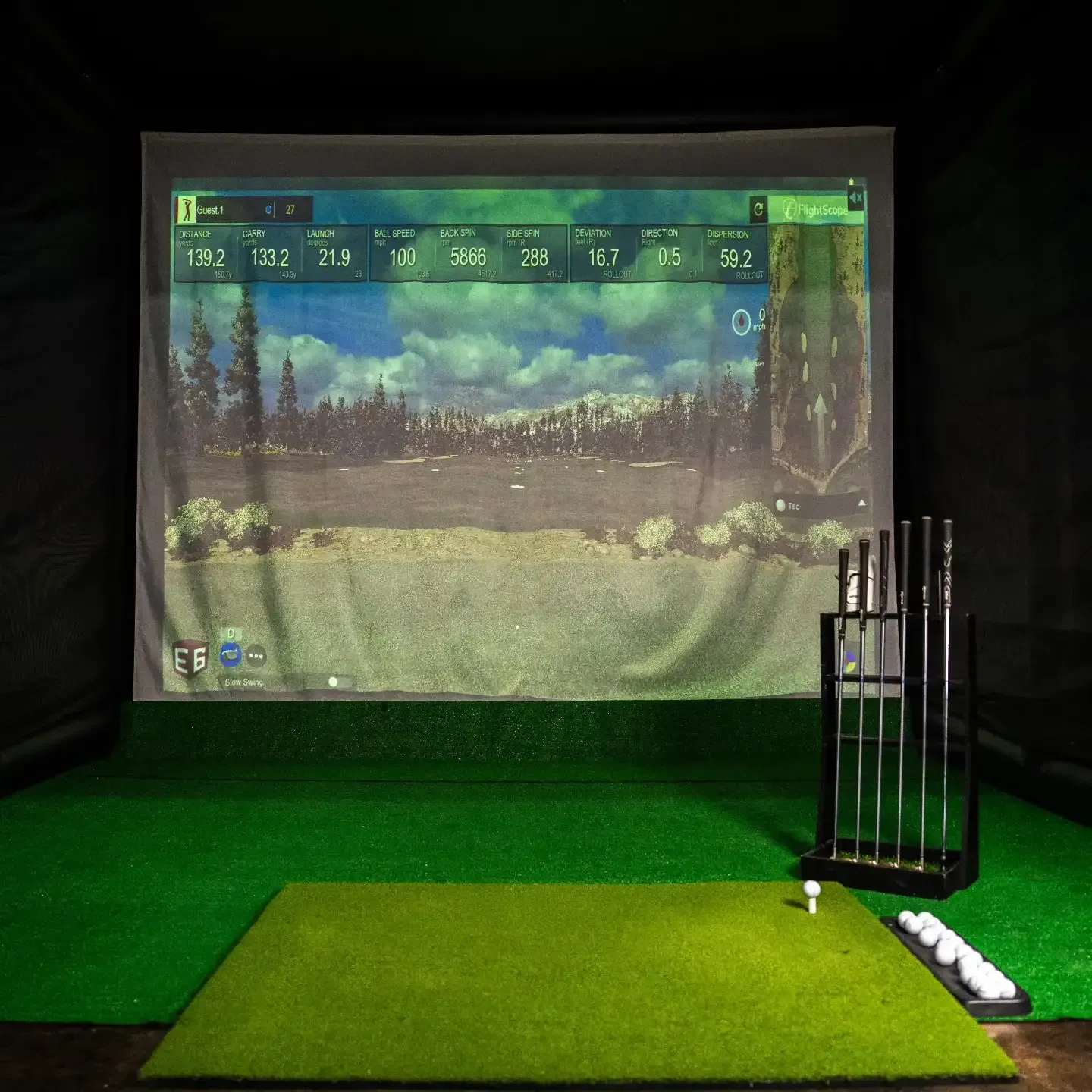 Indoor golf simulator with projected fairway, green hitting mat, and clubs beside a screen