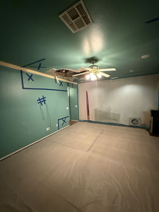 A room under construction: teal ceiling, covered floor, damaged ceiling with fan, partially painted walls.
